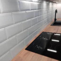Białe, błyszczące płytki ułożone w cegiełkę na ścianie nad blatem kuchennym z czarnym zlewem i płytą indukcyjną, oświetlenie LED pod szafkami.