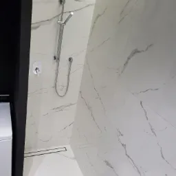 Kabina prysznicowa wykończona białymi płytkami imitującymi marmur, z chromowaną armaturą i odpływem liniowym w podłodze, widok z boku na fragment pralki.