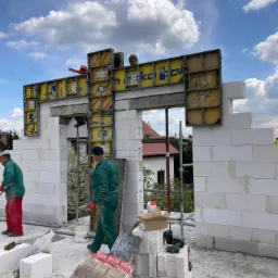 Wznoszenie ścian z białych bloczków, z użyciem żółtych szalunków Doka, dwóch robotników w ubraniach roboczych na placu budowy, częściowo widoczny dach sąsiedniego domu.