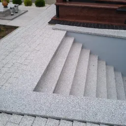 Granitowe schody prowadzące do piwnicy grobowca, otoczone kostką granitową i widocznym nagrobkiem w tle.