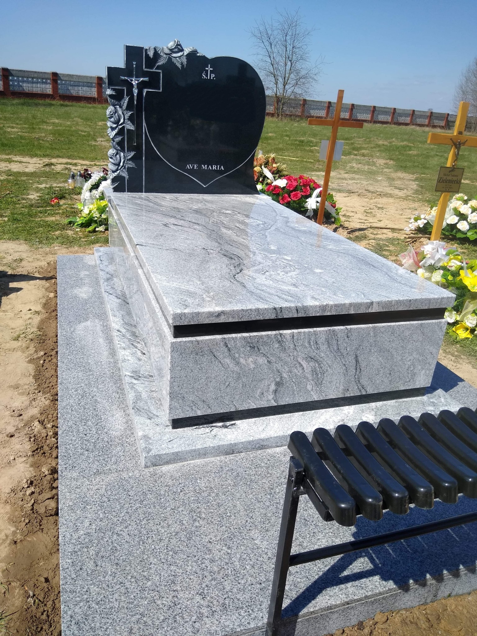 Granitowy nagrobek z czarnym, sercowatym elementem z krzyżem i różami, z napisem 'AVE MARIA', otoczony kwiatami i drewnianymi krzyżami na cmentarzu.