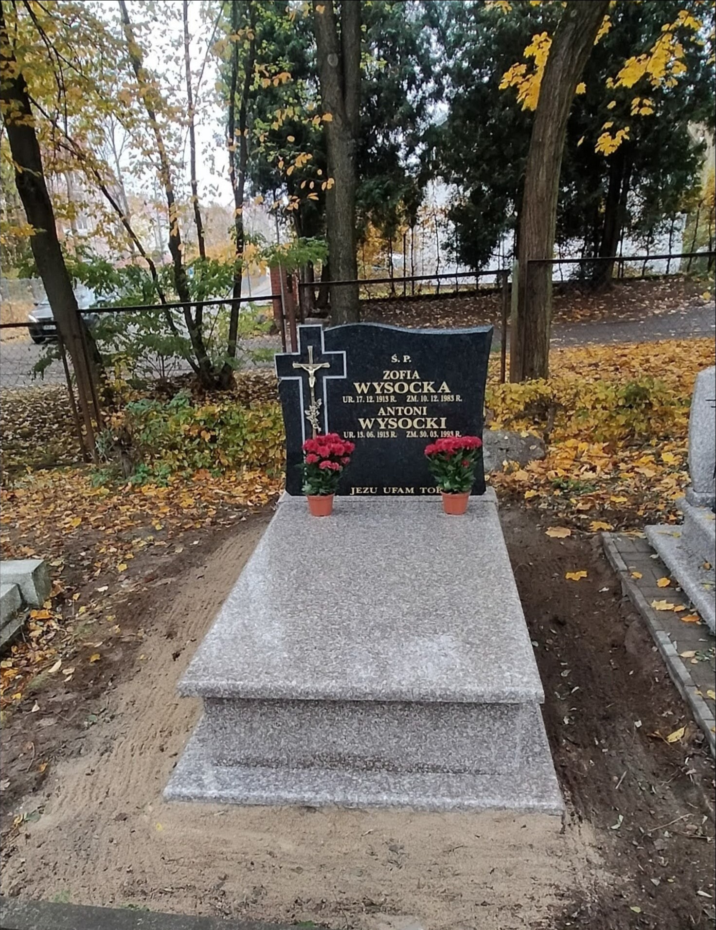 Granitowy nagrobek z czarną tablicą i krzyżem, ozdobiony dwoma doniczkami z czerwonymi kwiatami, na cmentarzu w Chorzelach, w jesiennej scenerii opadłych liści.