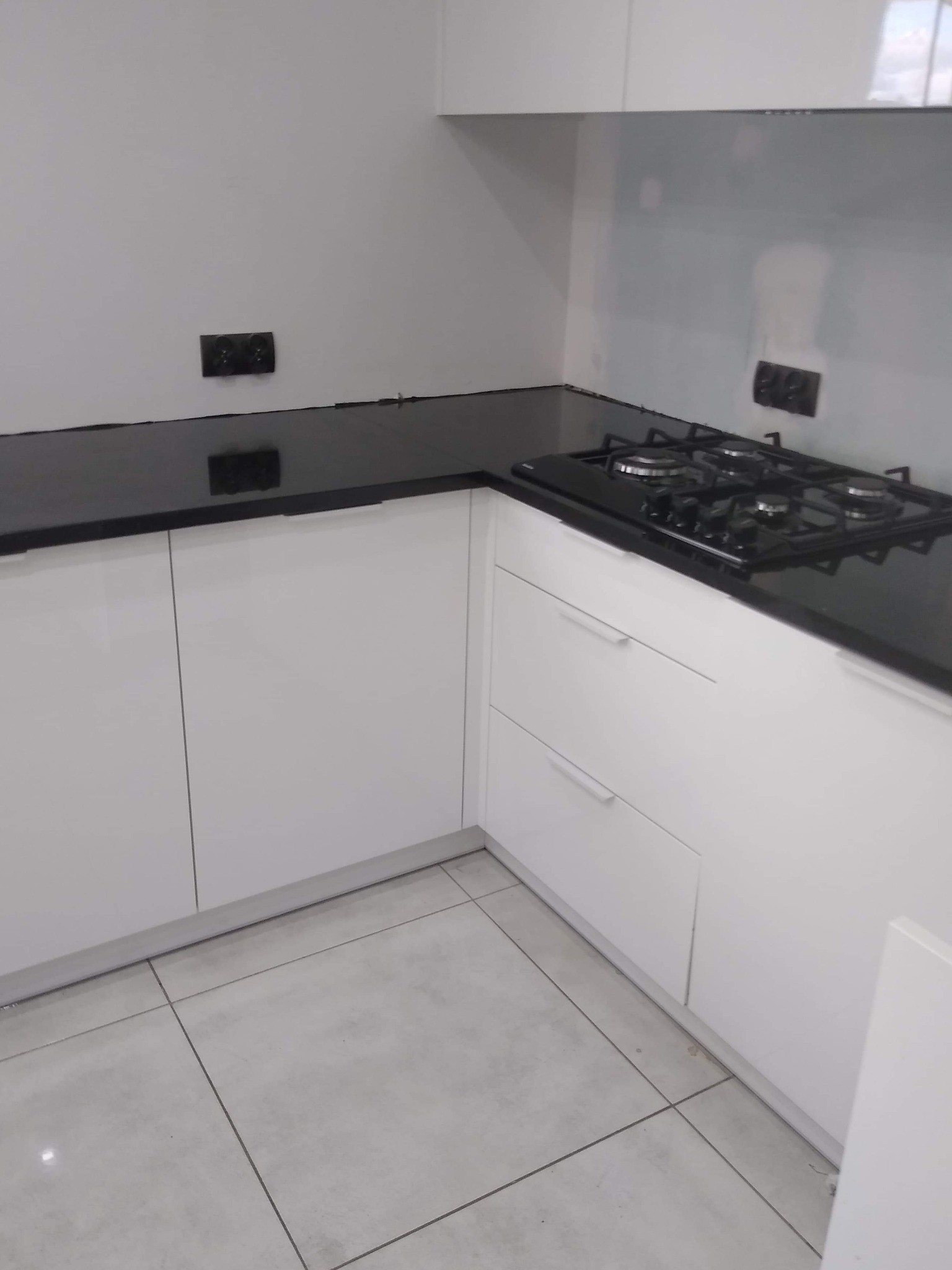 Biała, nowoczesna kuchnia z czarnym, błyszczącym blatem i płytą gazową, widok na fragment ściany z gniazdkami i białe płytki podłogowe.