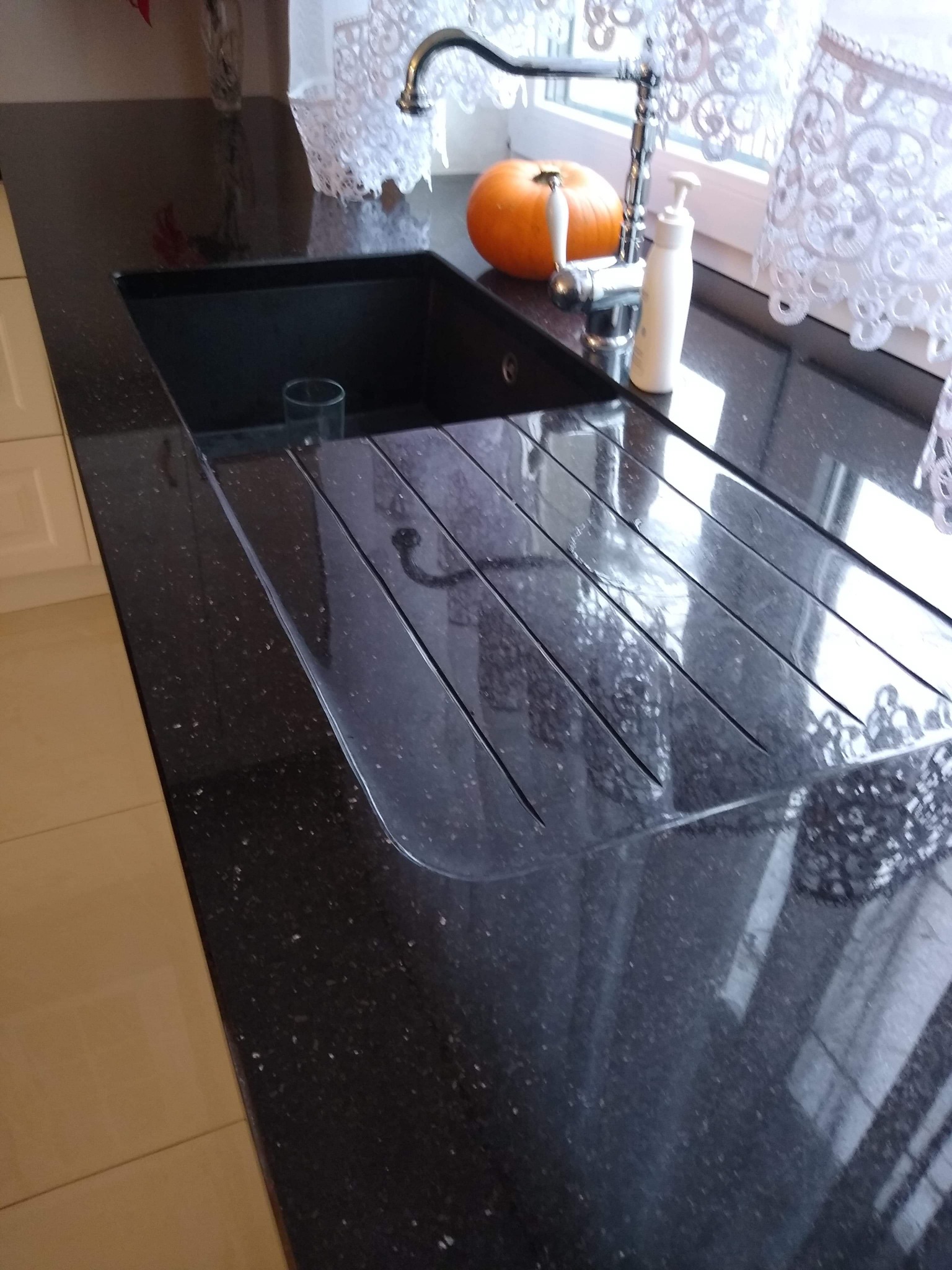 Czarny, błyszczący blat kuchenny z głębokim zlewem, ociekaczem z rowkami i dekoracyjną dynią oraz dozownikiem mydła przy kranie, na tle okna z koronkową firanką.