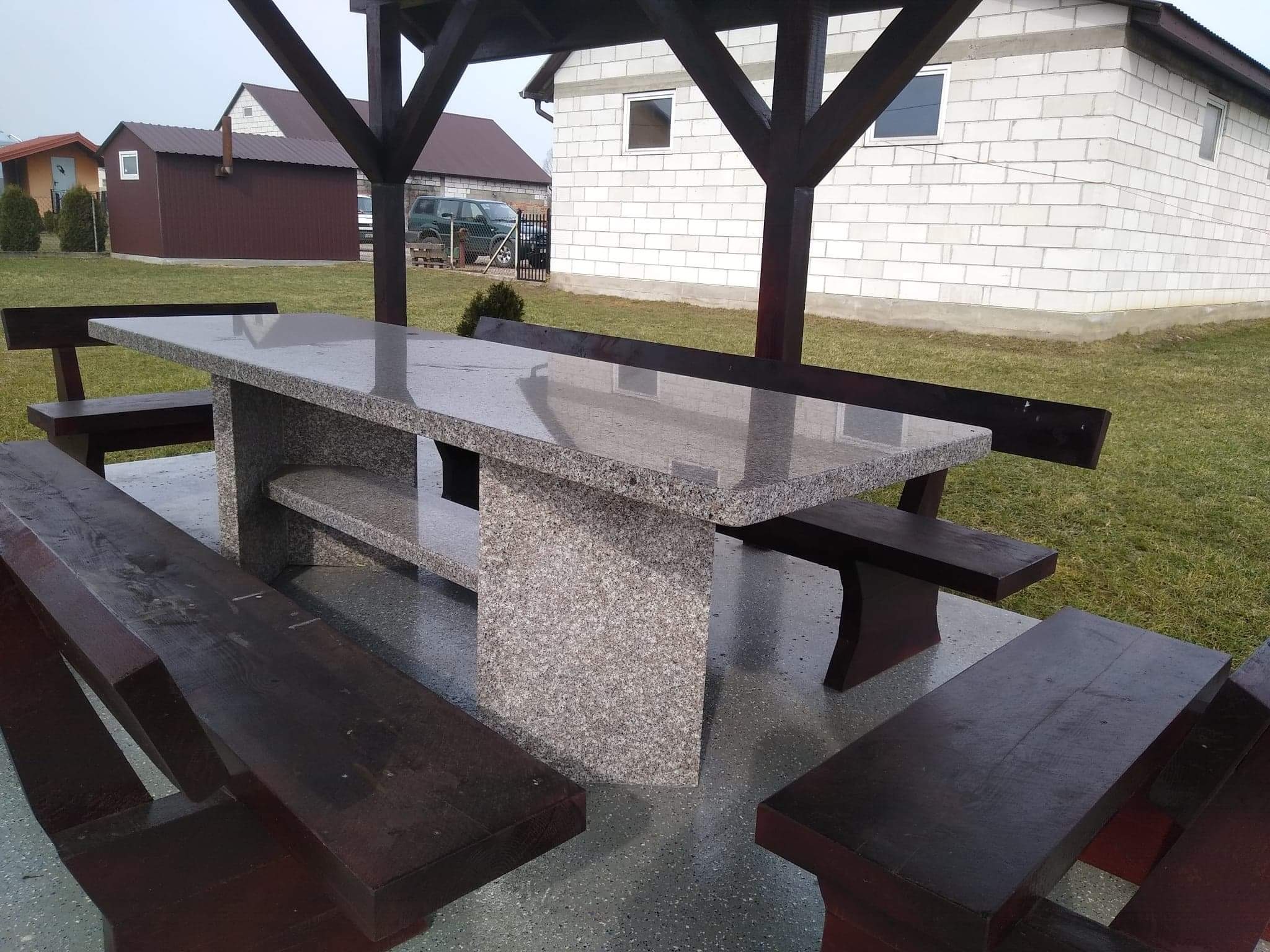 Zewnętrzny stół z blatem z szarego kamienia, zintegrowany z ławkami z ciemnego drewna, całość pod drewnianą konstrukcją zadaszenia, na zewnątrz budynki w tle.