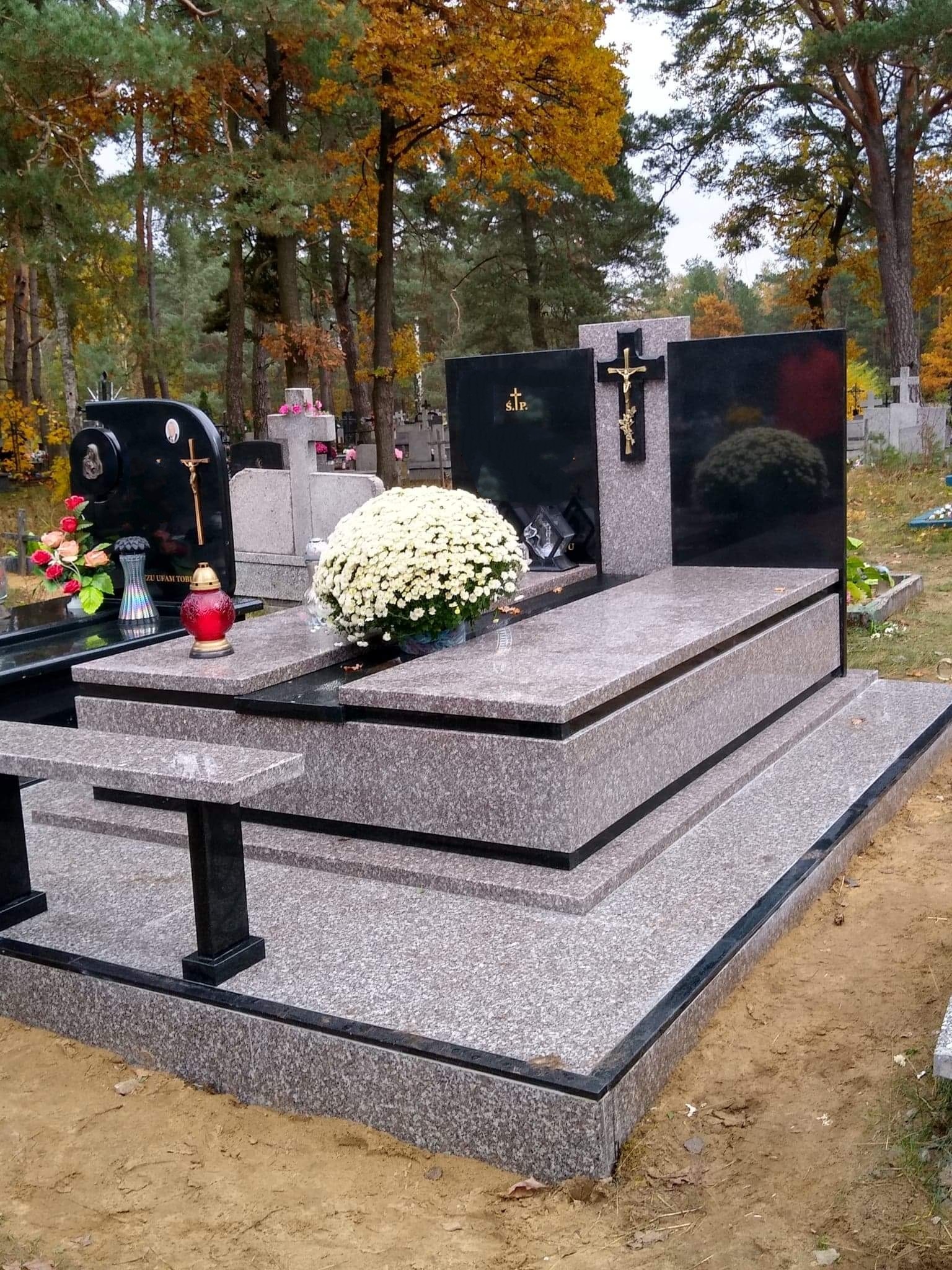 Granitowy nagrobek dwuosobowy z czarnymi tablicami, krzyżem i ławką, otoczony jesiennym krajobrazem cmentarza.