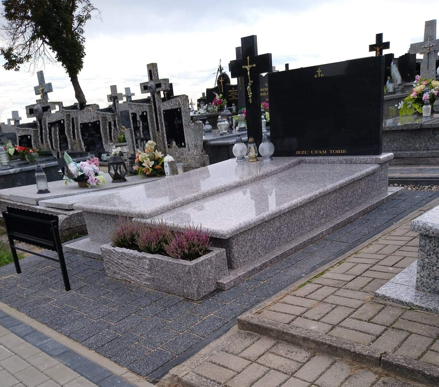 Granitowy nagrobek z czarną tablicą i napisem 'Jezu ufam Tobie', ozdobiony kwiatami wrzosu w kamiennej donicy, na tle innych nagrobków.