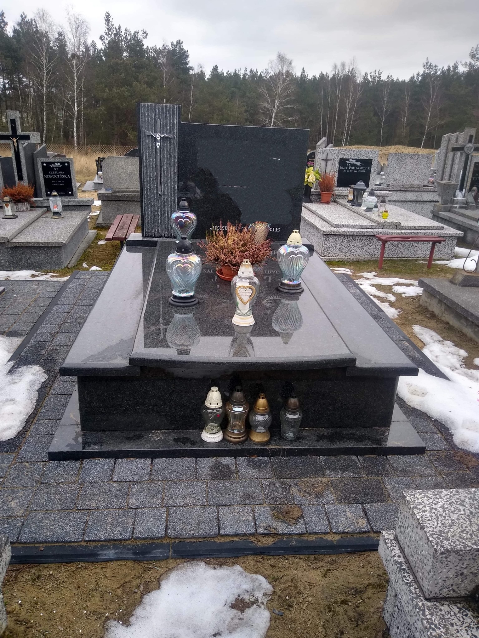 Czarny, dwupoziomowy pomnik z ciemnego granitu na cmentarzu, ozdobiony lampionami w kształcie serc i kwiatami w doniczce, otoczony kostką brukową i resztkami śniegu.