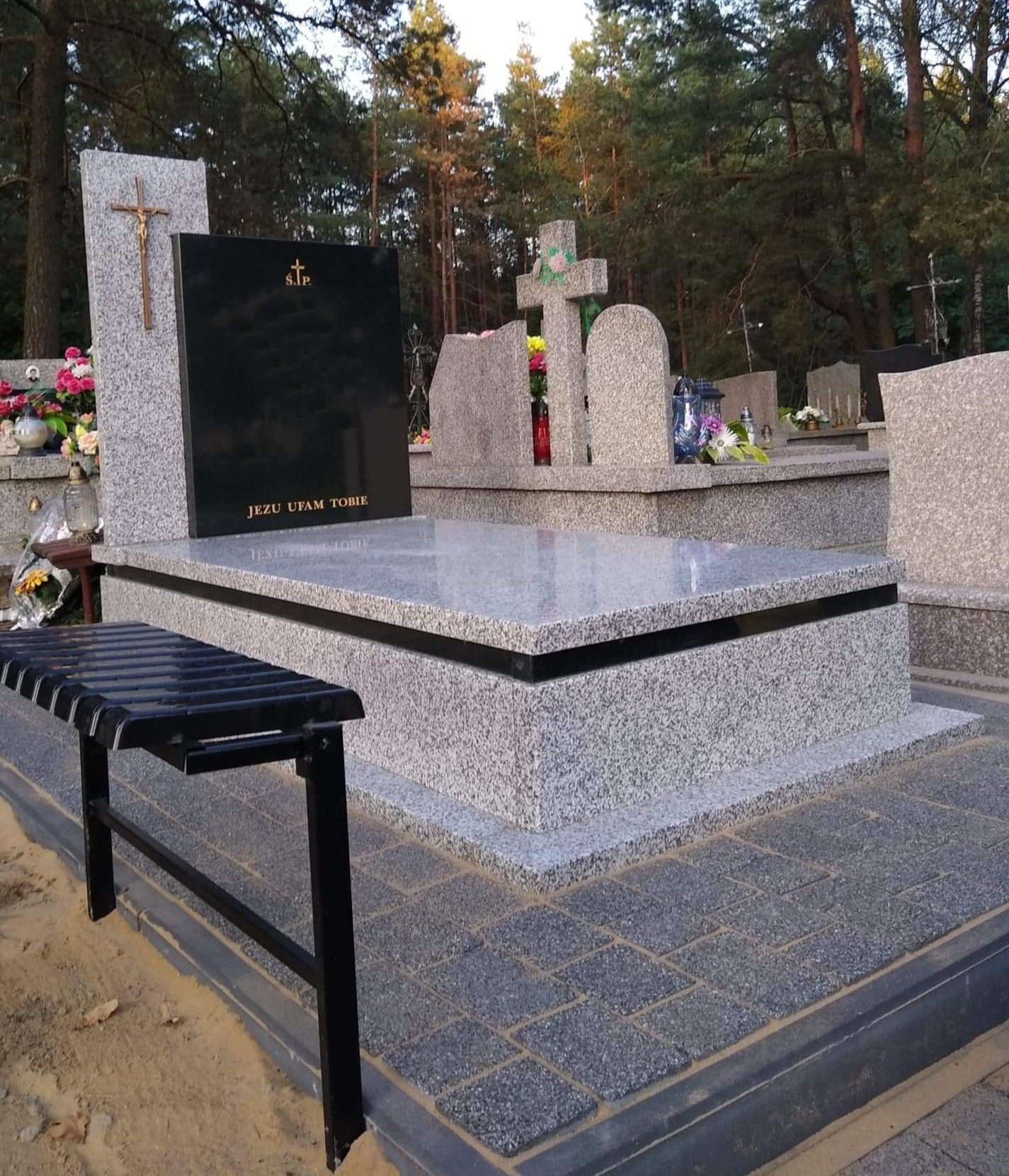 Granitowy pomnik z czarną tablicą i złotym krzyżem, napis 'Jezu Ufam Tobie', czarna metalowa ławeczka, kostka brukowa na cmentarzu, w tle drzewa.