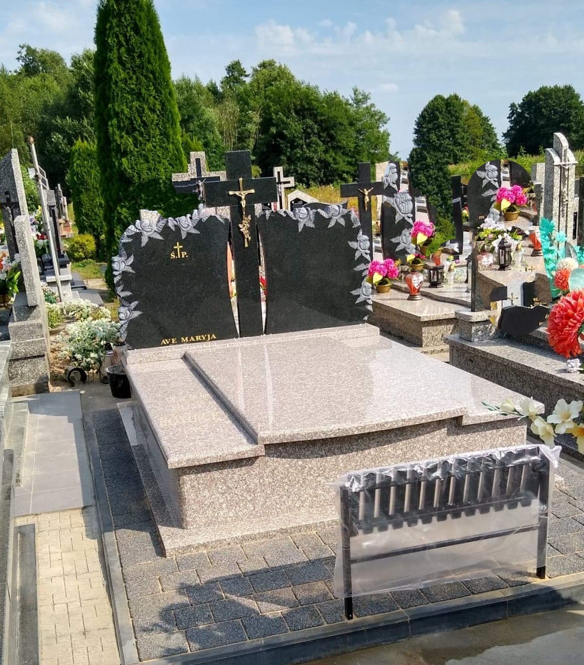 Podwójny, czarny nagrobek z jasnymi zdobieniami kwiatowymi i napisem 'AVE MARYJA', zwieńczony złotym krzyżem, na jasnym, kamiennym fundamencie, z widocznym stojakiem na znicze w folii, na cmentarzu.