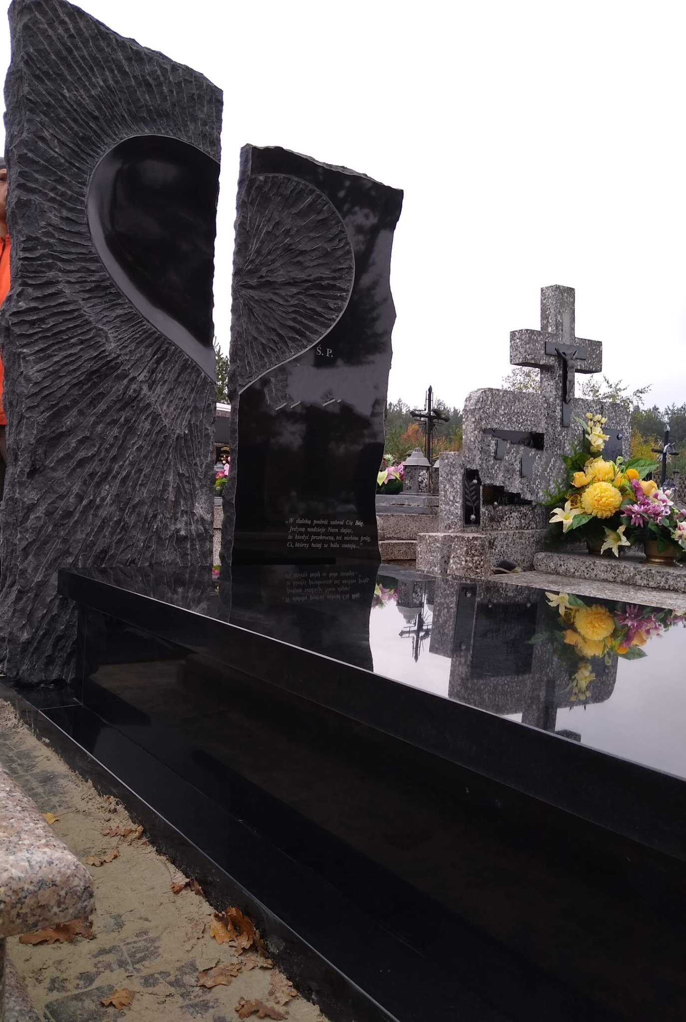 Czarny, nowoczesny nagrobek z elementem w kształcie serca i wyrytym tekstem, otoczony innymi nagrobkami na cmentarzu w pochmurny dzień.