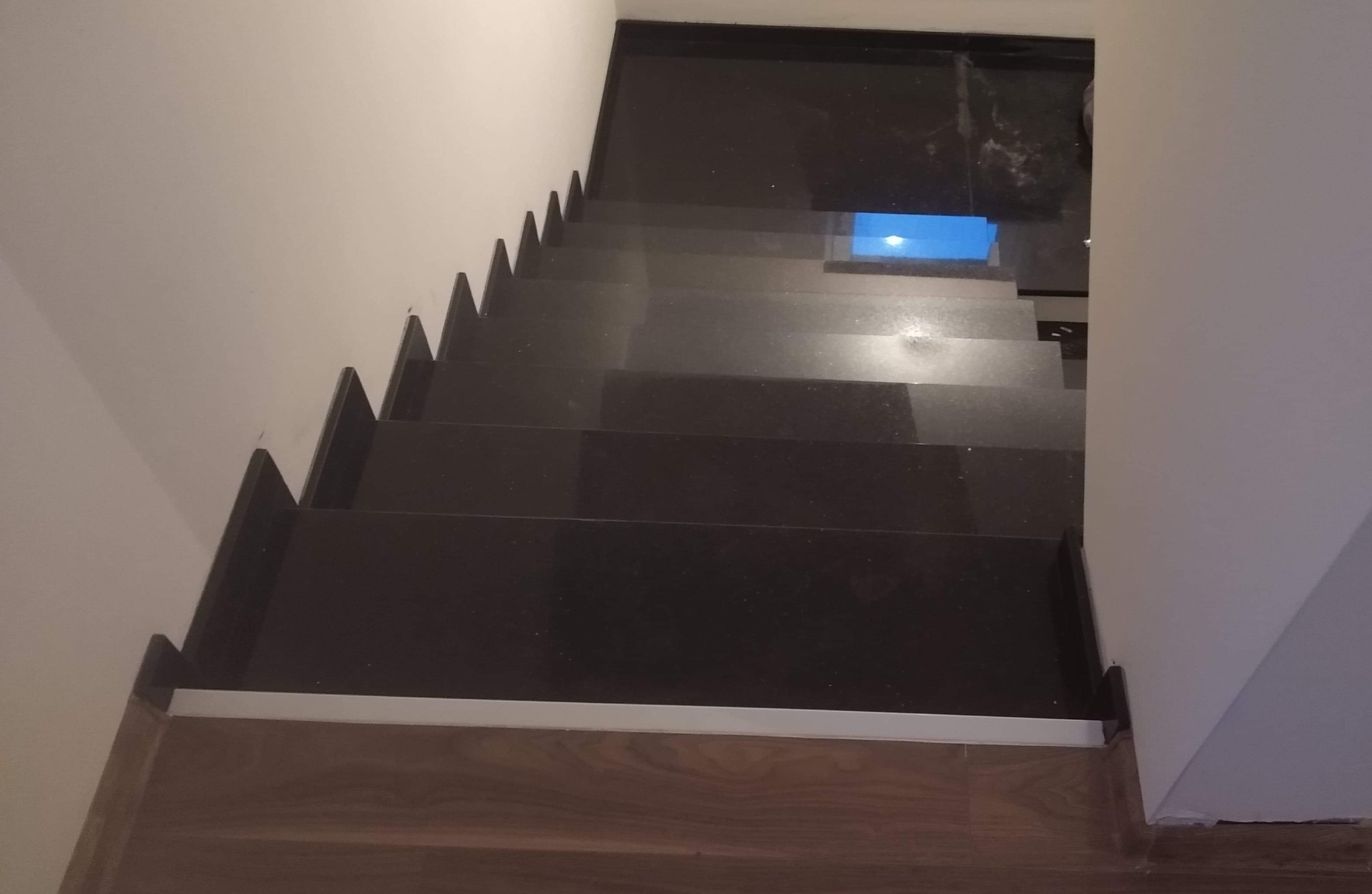 Ciemne, błyszczące schody z kamienia prowadzące w dół, widok z góry, oświetlone światłem wpadającym z otworu w suficie, z drewnianą podłogą u podstawy.