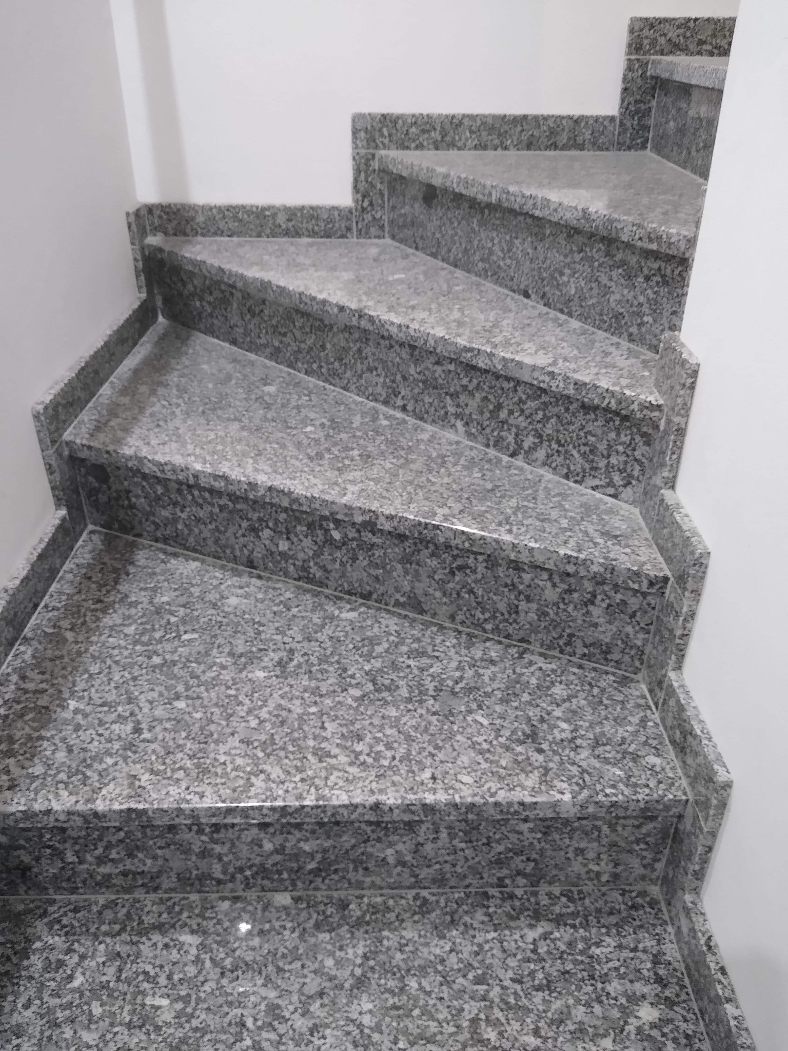 Granitowe schody wewnętrzne o szarej, nakrapianej fakturze, prowadzące wzdłuż białej ściany, widoczne z góry pod lekkim kątem.