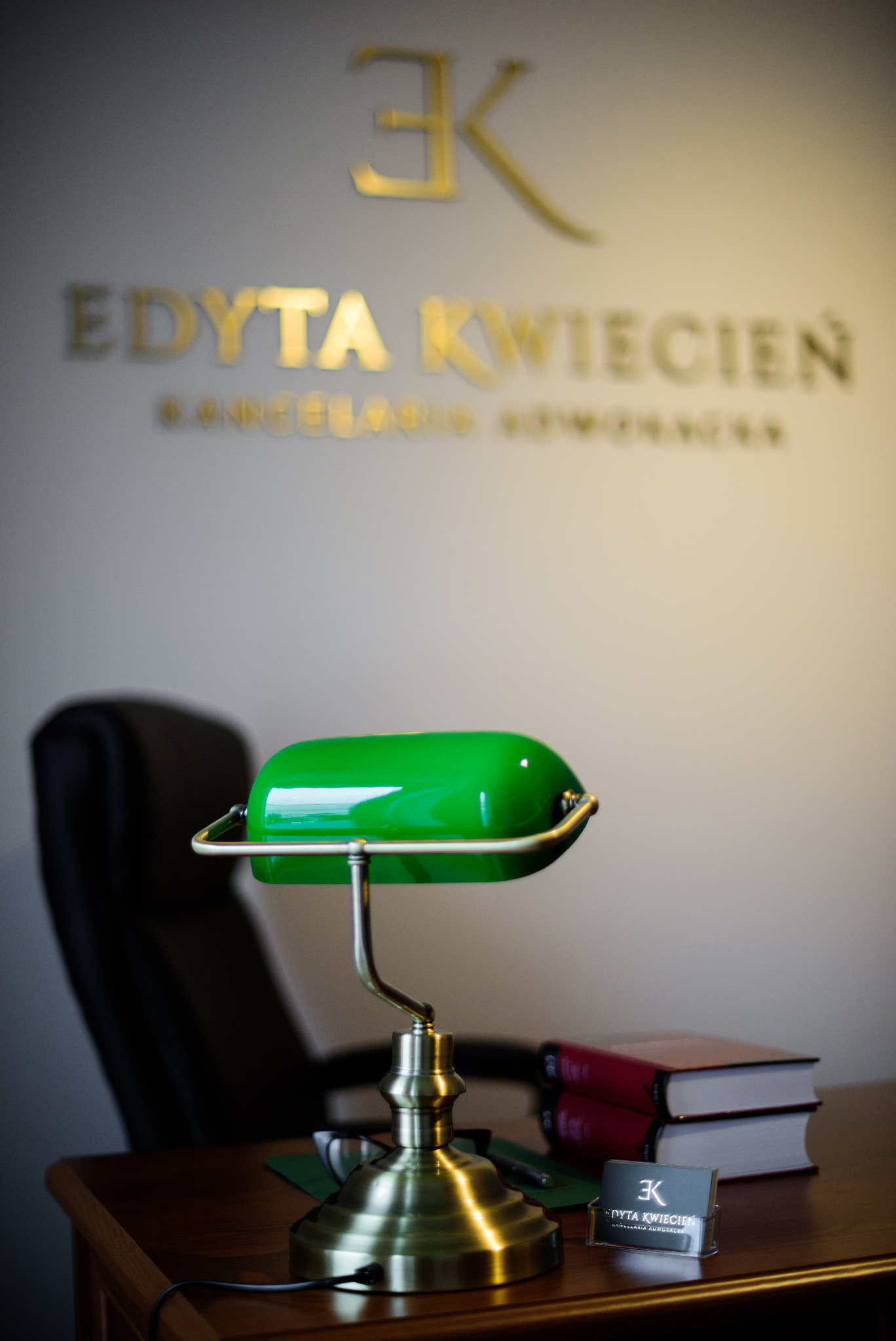 Gabinet z biurkiem, lampką bankierską, wizytówką i książkami, na ścianie logo 'EK' i nazwa 'Edyta Kwiecień Kancelaria Adwokacka'.