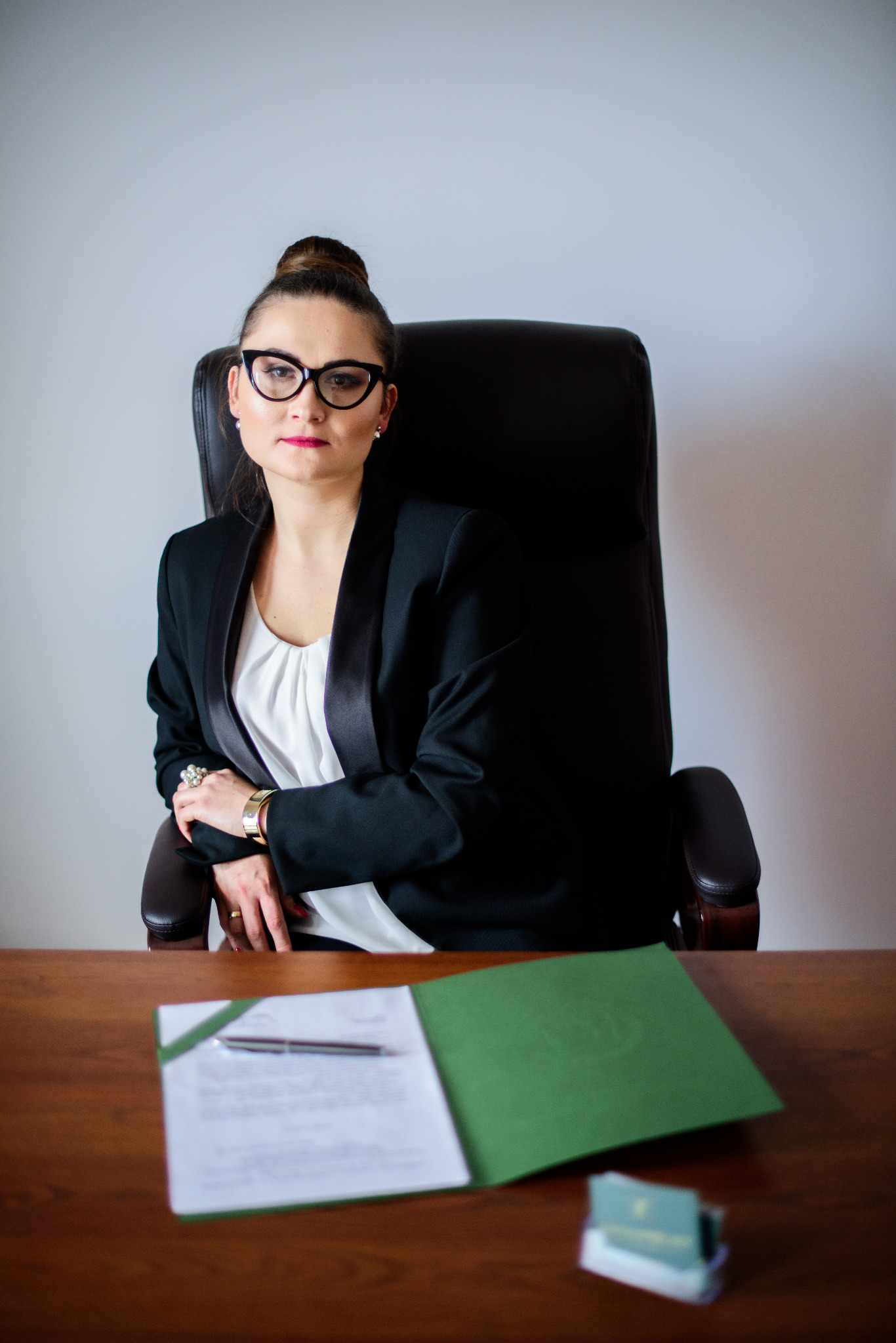 Kobieta w czarnej marynarce i okularach siedzi za biurkiem, na którym leży otwarty zielony segregator z dokumentami i długopisem.