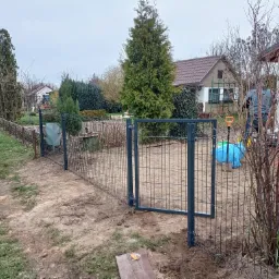 Montaż szarego ogrodzenia panelowego z bramką na posesji, widoczny fragment posesji, narzędzia i pracownik w tle.