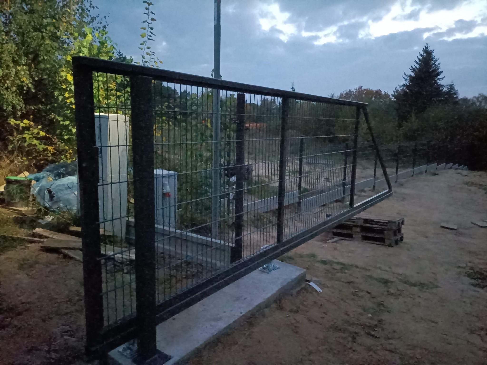 Suwana brama z metalowej siatki na betonowej podstawie, widok z boku na tle zachmurzonego nieba i zieleni, widoczne palety i elementy montażowe.