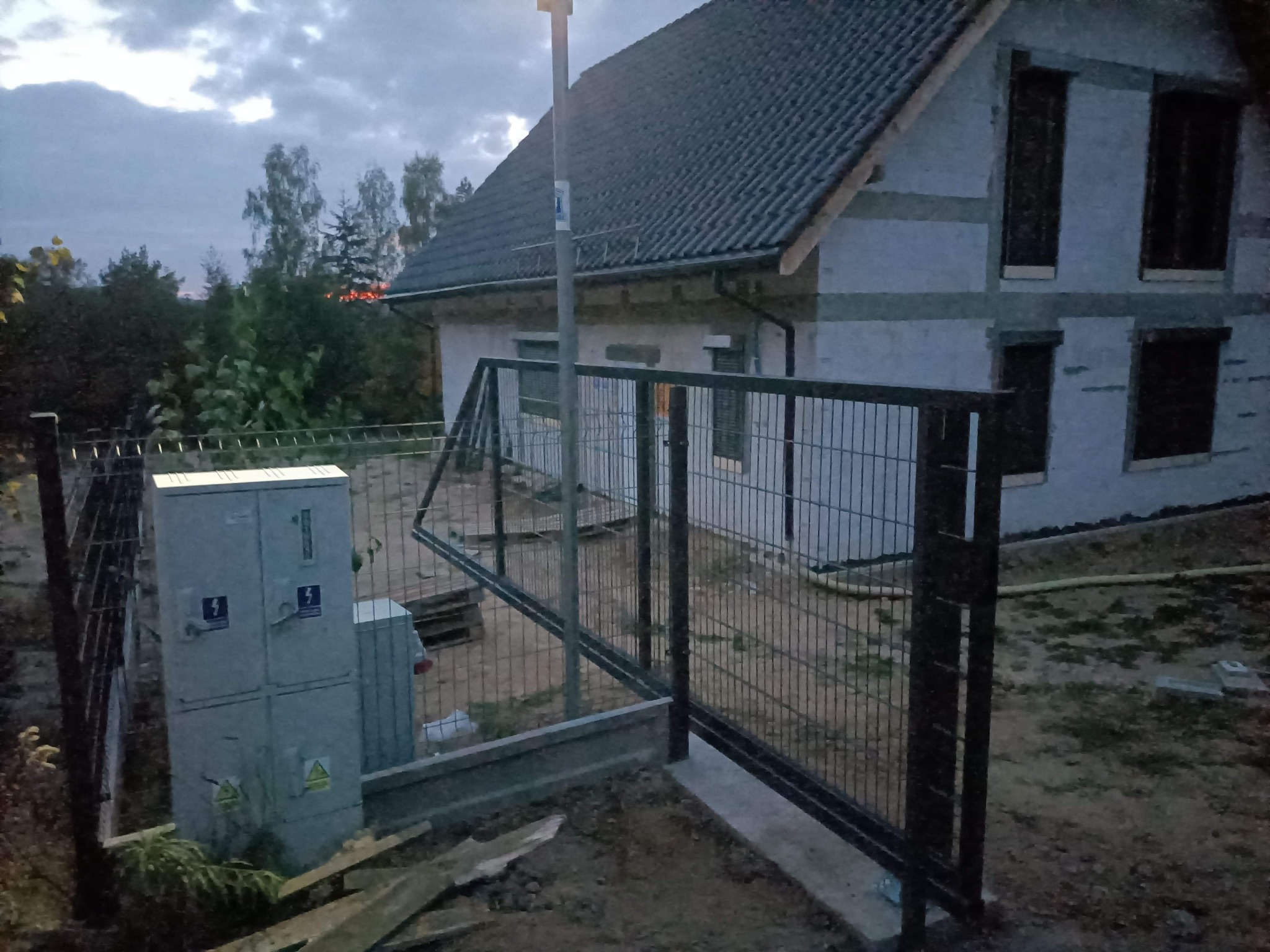 Nowoczesna, grafitowa brama panelowa z furtką, zamontowana przed domem w trakcie budowy, widoczna skrzynka elektryczna i lampa uliczna.