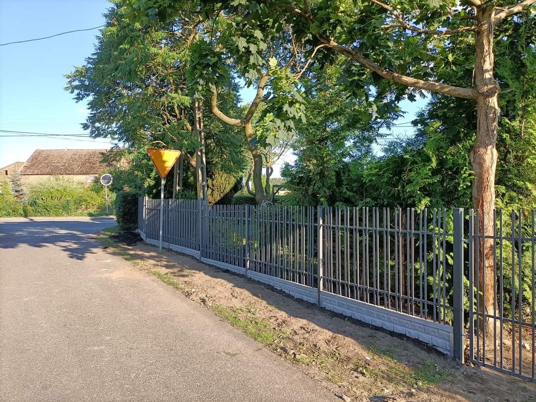Szare metalowe ogrodzenie ze słupkami i betonową podmurówką, widok z poziomu ulicy, w tle zieleń i dom z brązowym dachem.