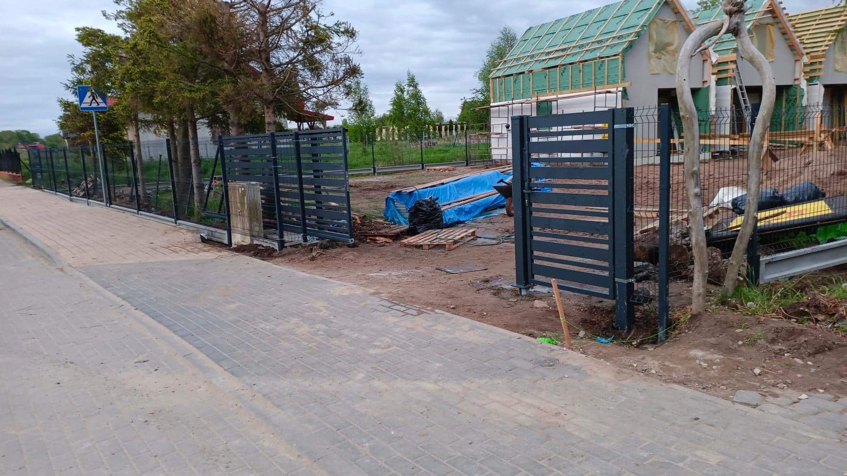 Montaż nowoczesnego ogrodzenia panelowego z bramą wjazdową, widoczne prace ziemne i przygotowanie terenu, w tle budynki w trakcie budowy.