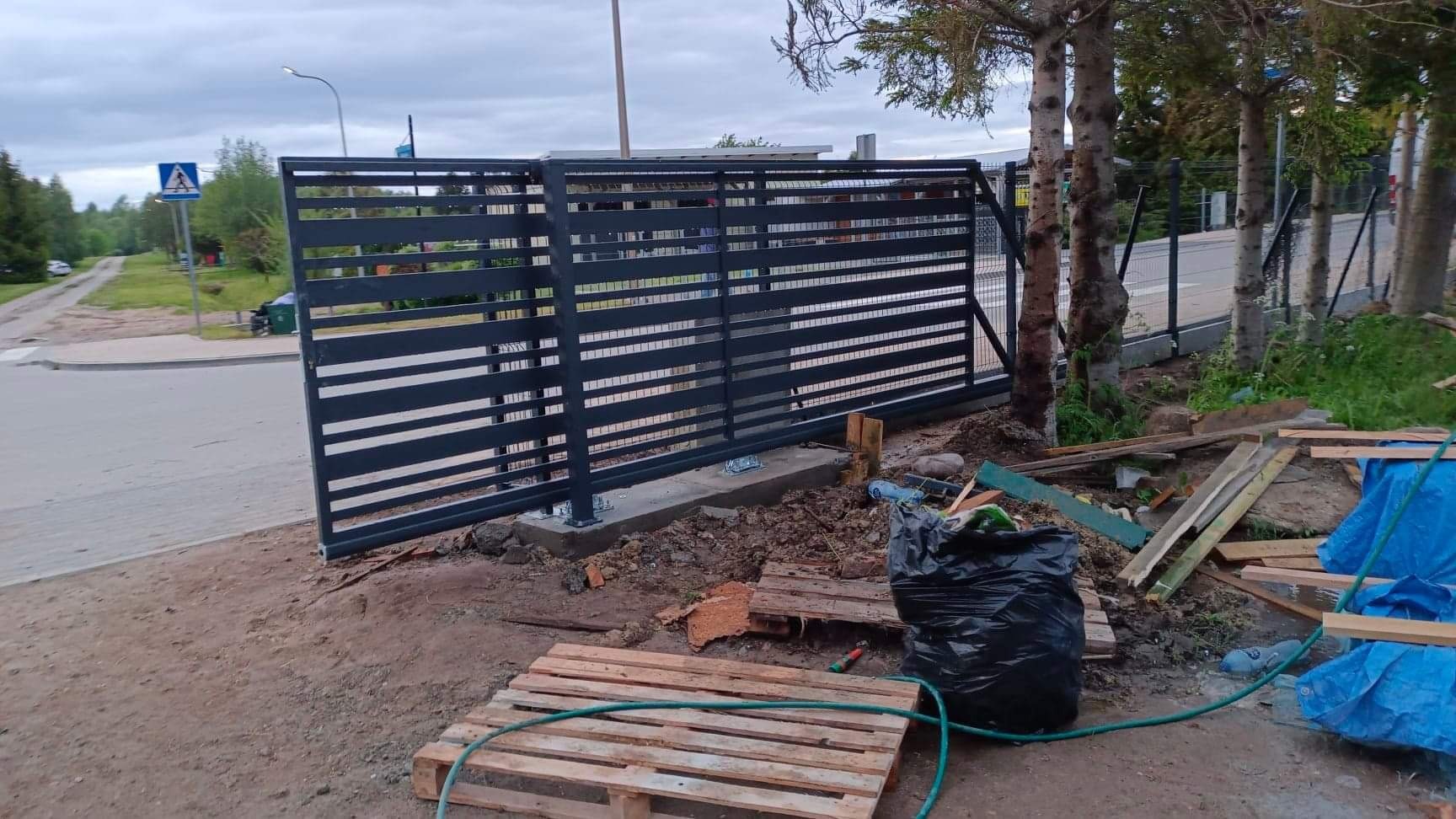 Suwana brama wjazdowa z poziomymi profilami, widoczna w trakcie montażu, z elementami rusztowania i workiem na śmieci w tle.