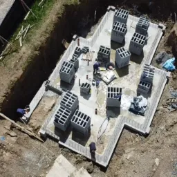 Widok z góry na plac budowy z wykopanym fundamentem, częściowo zalanym betonem i ułożonymi pustakami szalunkowymi; widoczny mały żółty koparka i pracownicy budowlani.