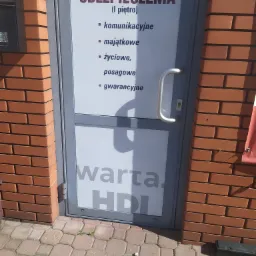 Wejście do biura Warta HDI z informacją o ubezpieczeniach komunikacyjnych, majątkowych, życiowych, posagowych i gwarancyjnych, widoczne z zewnątrz, obok ceglana ściana.