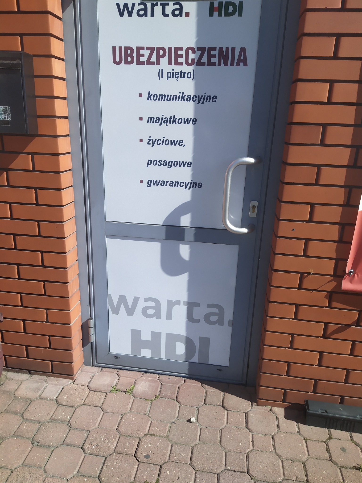 Wejście do biura Warta HDI z informacją o ubezpieczeniach komunikacyjnych, majątkowych, życiowych, posagowych i gwarancyjnych, widoczne z zewnątrz, obok ceglana ściana.