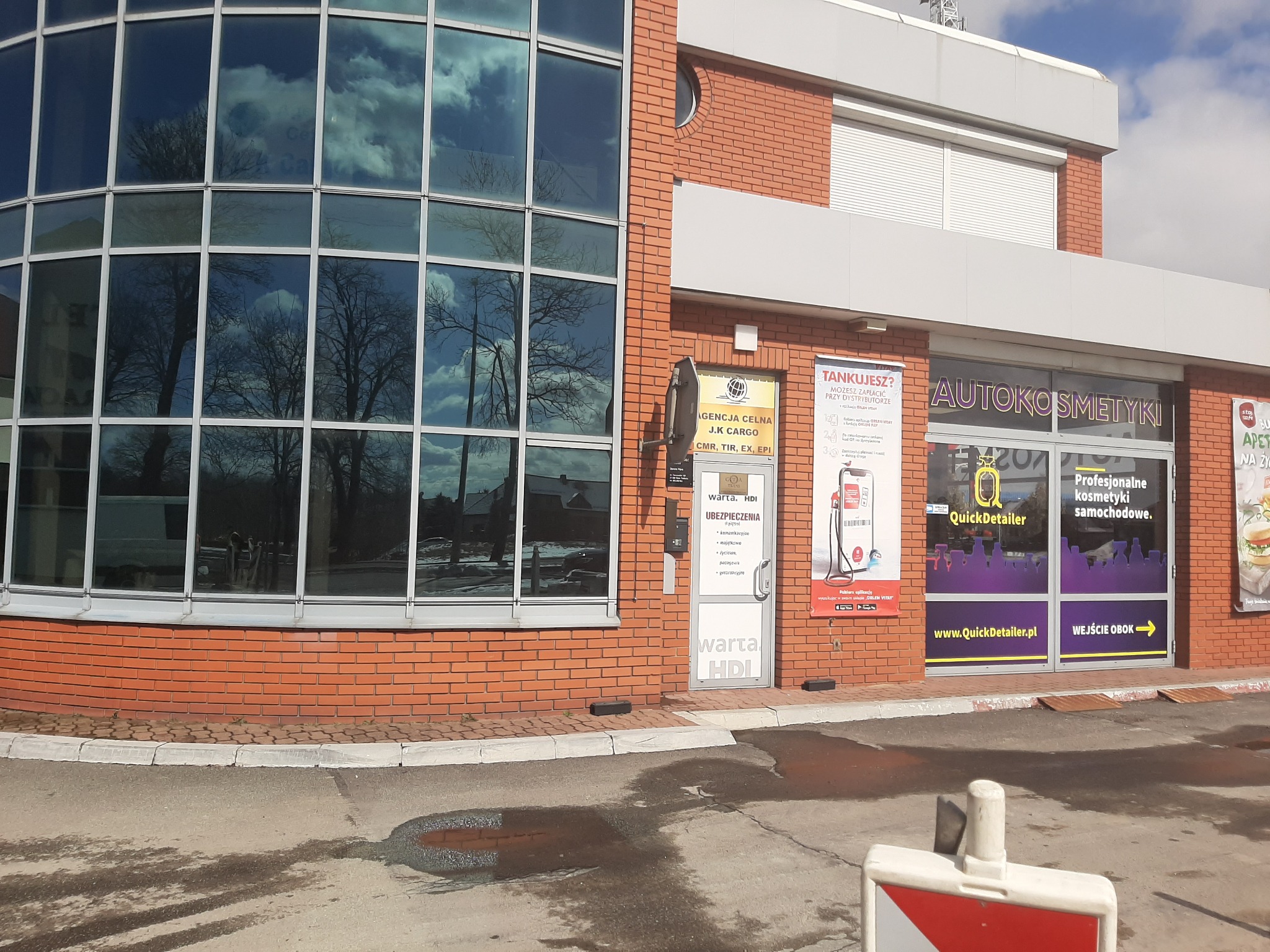 Budynek agencji celnej i punktu ubezpieczeń Warta HDI z widoczną ofertą ubezpieczeń majątkowych, komunikacyjnych, na życie i podróżnych, obok autokosmetyki QuickDetailer z wejściem obok.