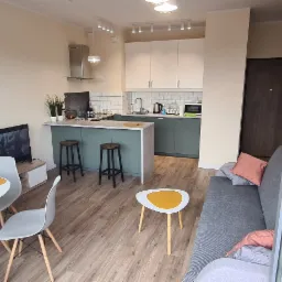 Nowoczesne wnętrze mieszkania z otwartą kuchnią, widok na stół z krzesłami, barek z hokerami i sofę przy oknie z odbiciem panoramy miasta.