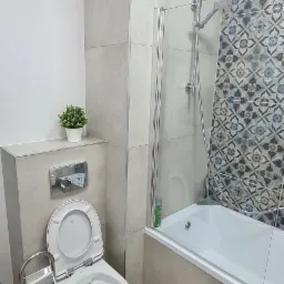 Nowoczesna łazienka z szarymi płytkami imitującymi beton, białą wanną z parawanem nawannowym, wiszącą toaletą z otwartą deską, chromowanym zestawem WC i małą rośliną doniczkową.