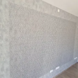 Ściana w pokoju po remoncie z dekoracyjną tapetą o geometrycznym wzorze, obok ściana pomalowana farbą w odcieniach szarości z widocznymi gniazdkami elektrycznymi i włącznikiem światła.