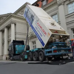 W Dębem Wielkim, ciężarówka AlfaFloor z podnośnikiem i zbiornikiem na wylewkę anhydrytową, z operatorem w kasku, na tle budynku z kolumnami podczas wylewania materiału na czarną matę ochronną.