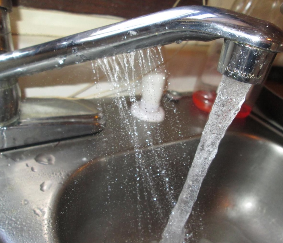 Woda lejąca się z chromowanego kranu do metalowego zlewu, widoczne drobne krople wody i odbicia światła.