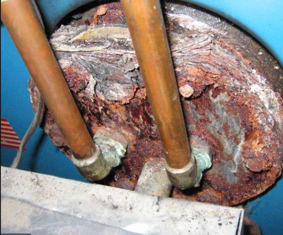 Silnie skorodowany element instalacji hydraulicznej z miedzianymi rurami, widoczne ślady rdzy i osadów, zielonkawy nalot na łączeniach, niebieskie tło zbiornika.