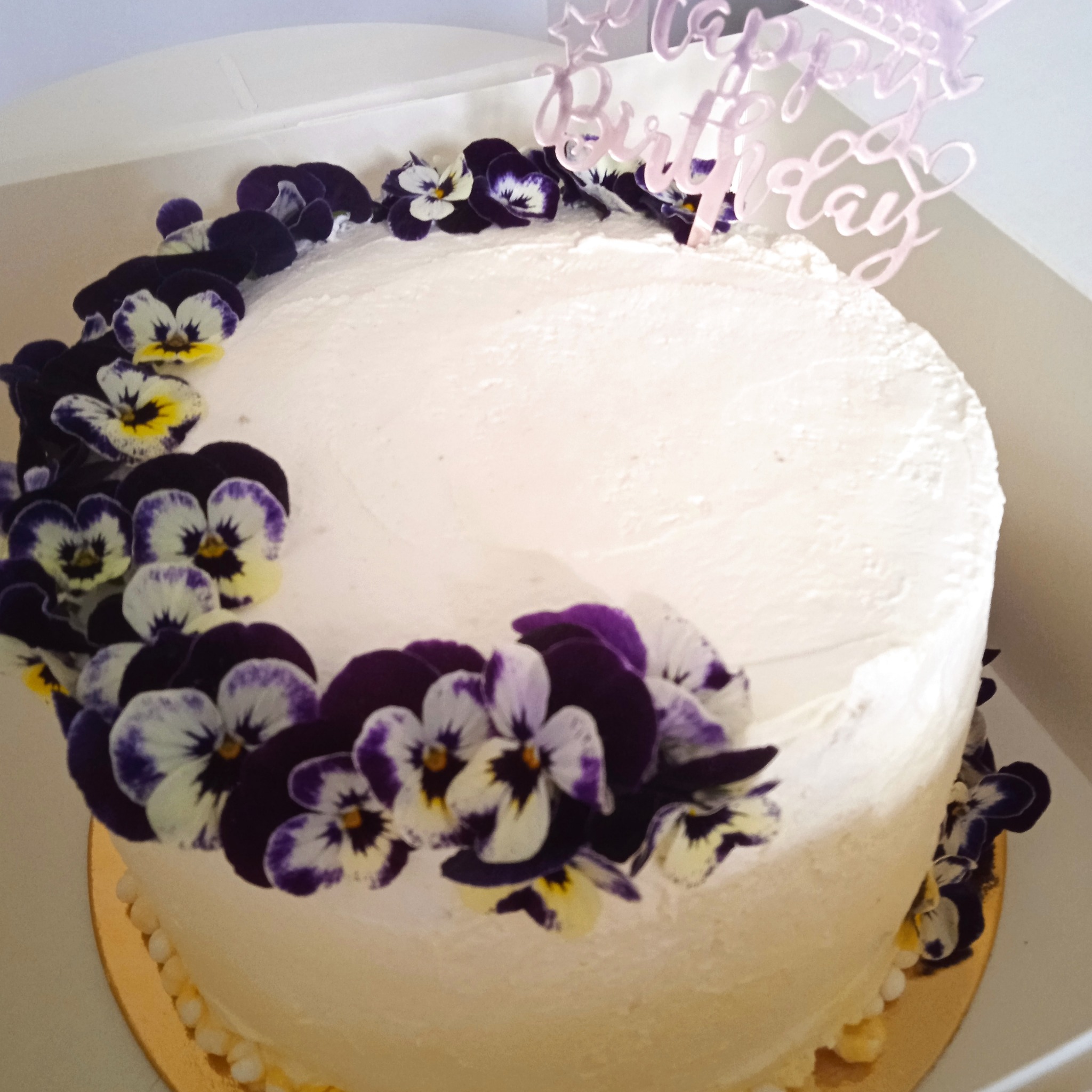 Tort urodzinowy z białym kremem, udekorowany fioletowymi i białymi bratkami oraz topperem 'Happy Birthday', umieszczony w białym pudełku.