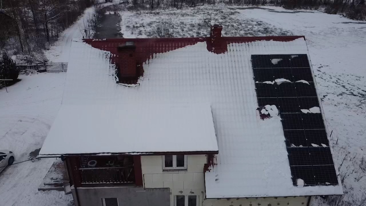 Zimowy widok z góry na dom z czerwonym dachem i panelami słonecznymi częściowo zasypanymi śniegiem, w tle ośnieżone drzewa.