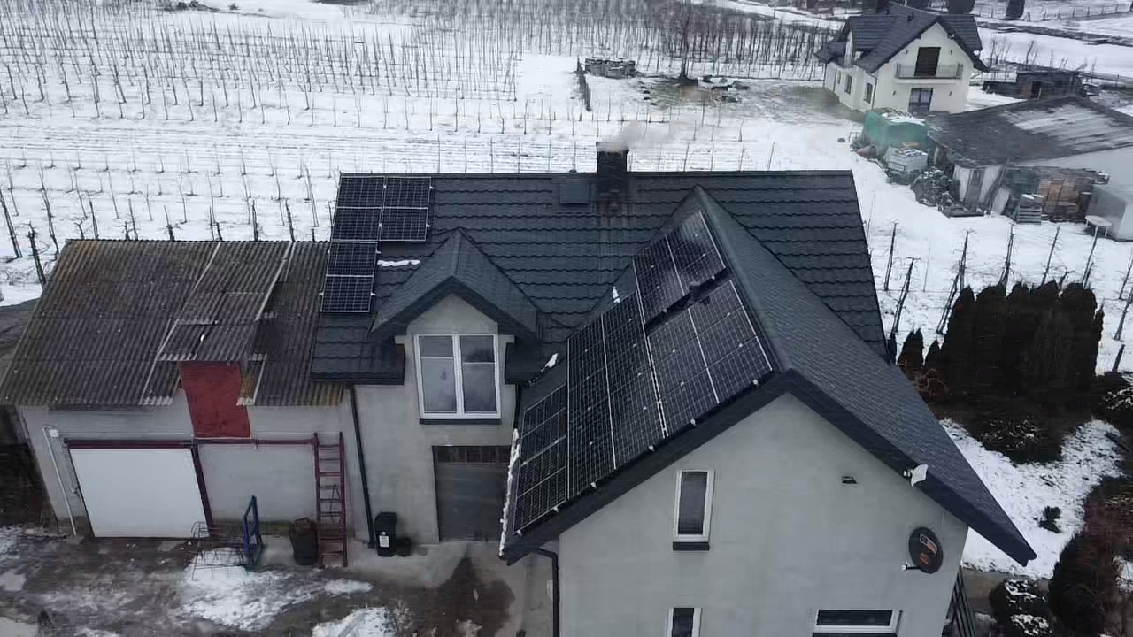 Widok z góry na dom z panelami słonecznymi na dachu, komin dymi, zimowy krajobraz w tle.
