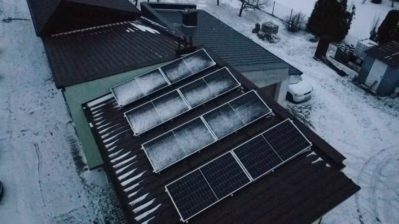Zimowy widok z góry na panele fotowoltaiczne pokryte śniegiem na ciemnym dachu domu jednorodzinnego, z zasypanym śniegiem podjazdem i samochodem w tle.