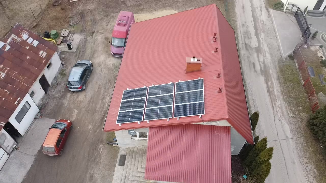 Widok z góry na dom z czerwonym dachem, na którym zamontowane są trzy panele fotowoltaiczne; obok domu zaparkowane samochody.