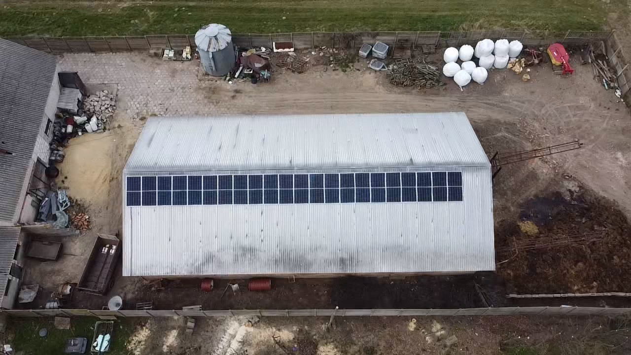 Widok z drona na duży budynek gospodarczy z zamontowanymi panelami słonecznymi na dachu, otoczony ziemią i innymi budynkami gospodarczymi.