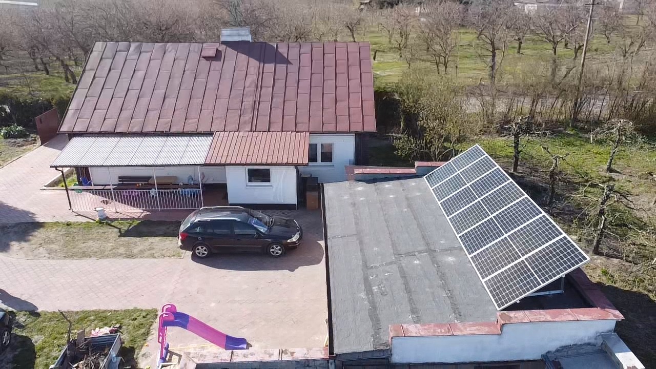 Widok z góry na dom z panelami słonecznymi zamontowanymi na płaskim dachu, obok zaparkowany samochód osobowy i zjeżdżalnia dla dzieci.