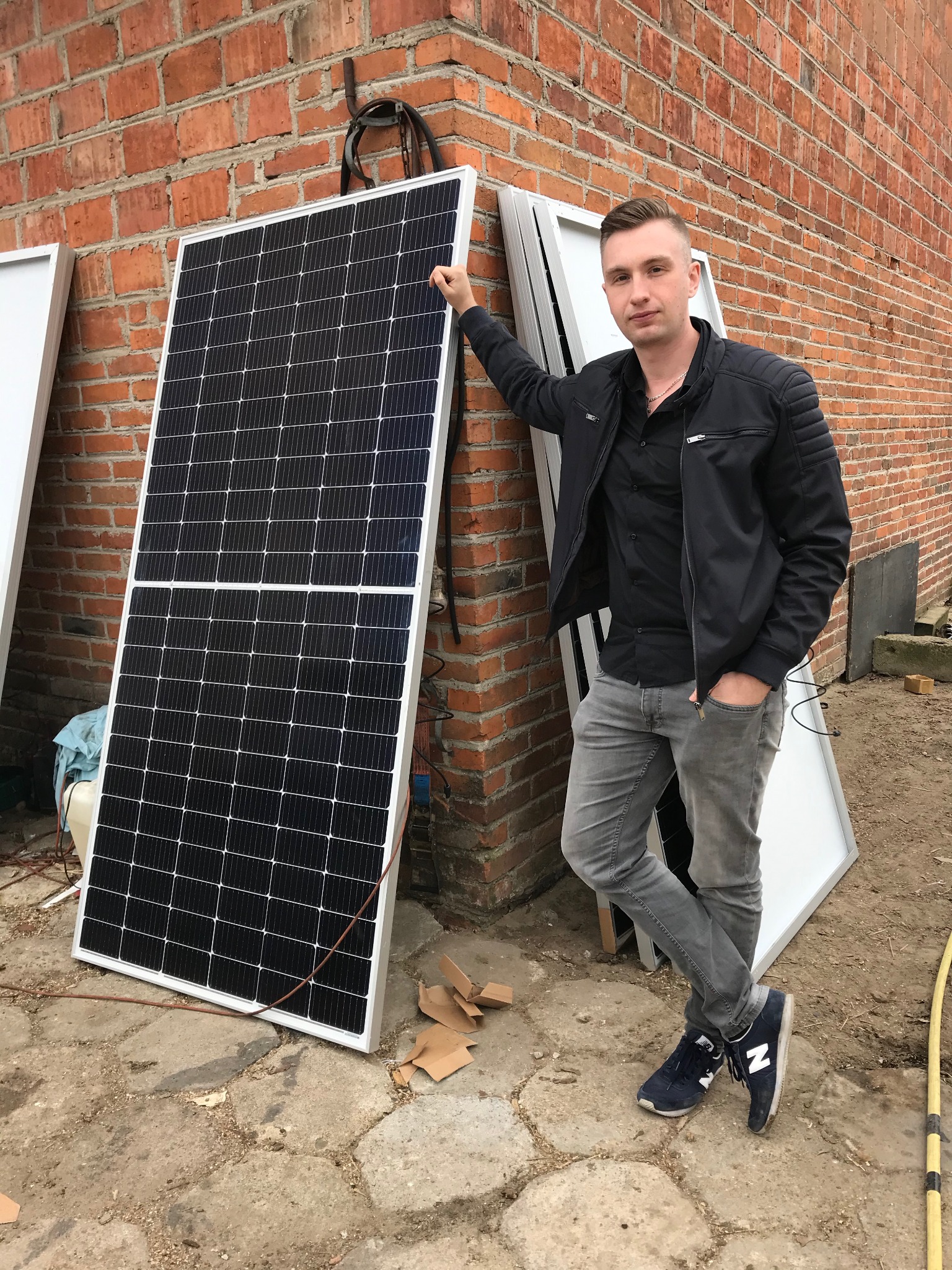 Mężczyzna w czarnej kurtce opiera się o panel słoneczny na tle ceglanej ściany, obok leżą dodatkowe panele.