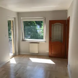 Jasne, puste pomieszczenie z parkietem w jodełkę, świeżo pomalowanymi szarymi ścianami, oknem z widokiem na zieleń i drewnianymi drzwiami z przeszkleniami.