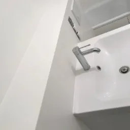 Biała, nowoczesna umywalka z chromowaną baterią i odpływem, zamontowana w minimalistycznej łazience z białymi płytkami i ukrytym systemem spłukiwania toalety.