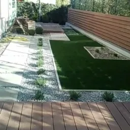 Nowoczesny ogród z drewnianym tarasem, kamienną ścieżką, równo przyciętym trawnikiem i drewnianym ogrodzeniem.