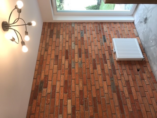 Ściana z cegły klinkierowej w odcieniach czerwieni i brązu, oświetlona lampą sufitową z żarówkami Edison, z widocznym grzejnikiem i oknem dachowym.