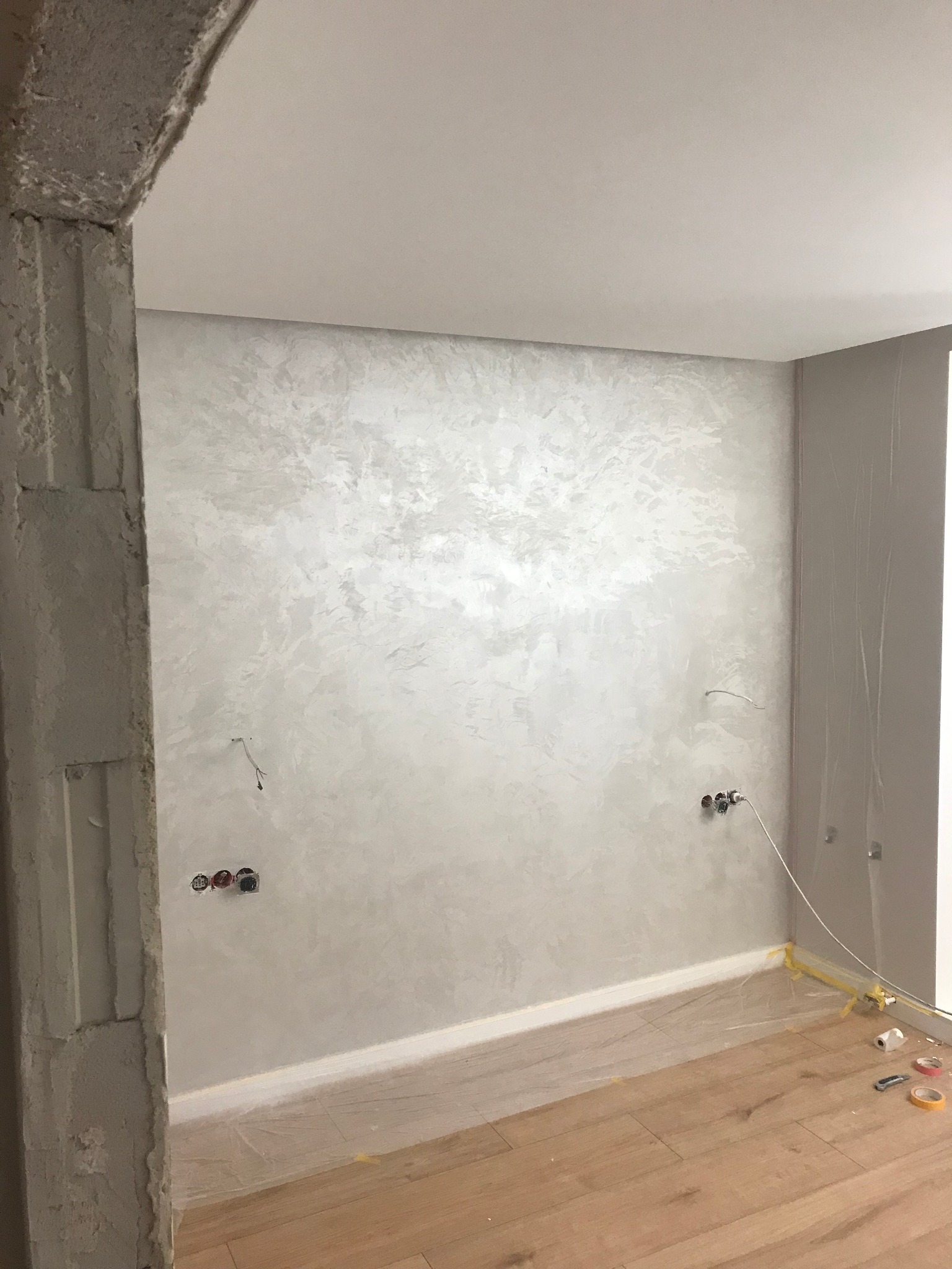 Ściana wykończona dekoracyjnym tynkiem w odcieniach szarości, z widocznymi gniazdkami elektrycznymi i zabezpieczoną podłogą drewnianą folią malarską oraz taśmą, fragment surowej ościeżnicy po lewej...