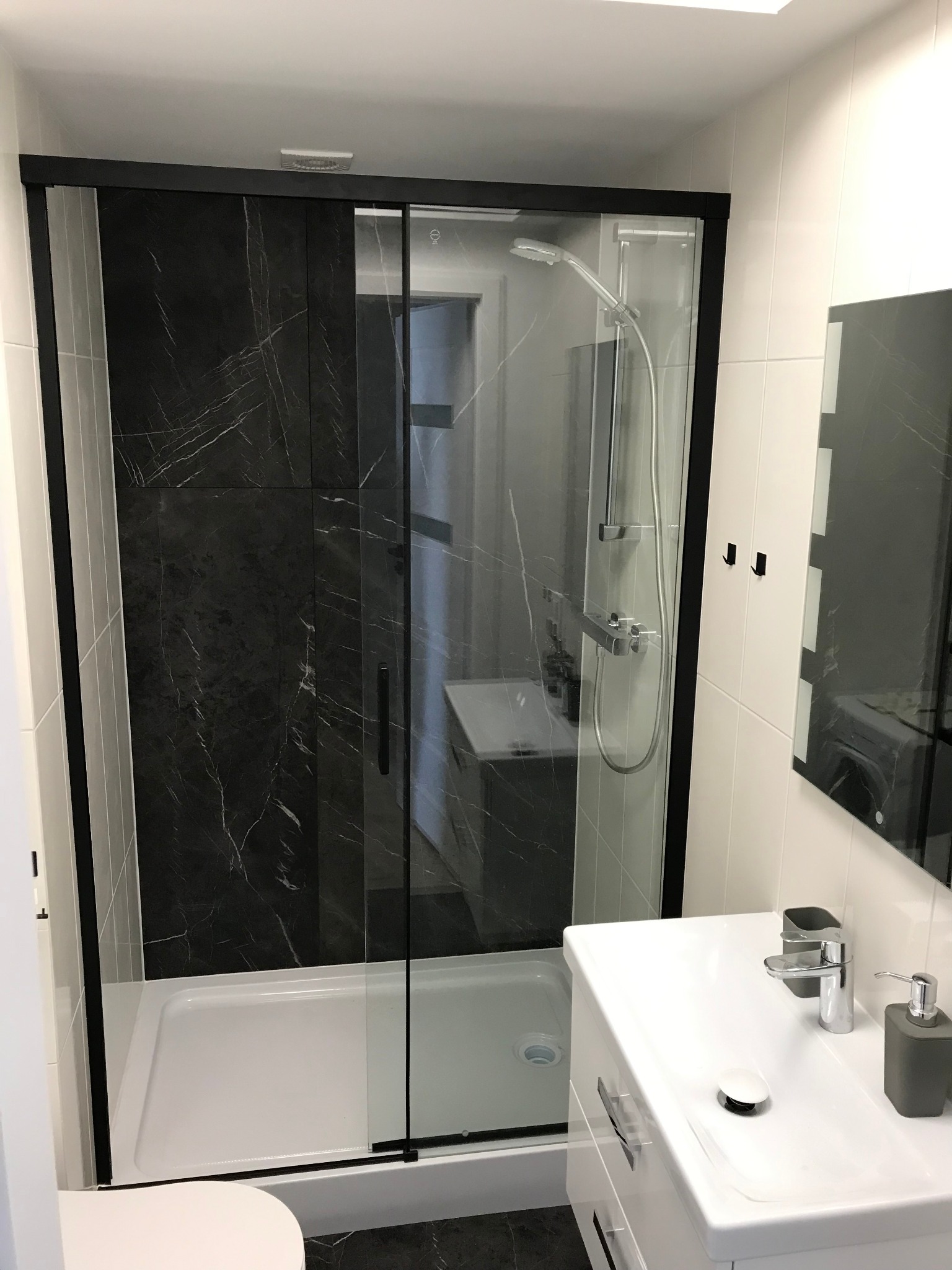 Nowoczesna łazienka z kabiną prysznicową z czarnymi ramami i panelami imitującymi marmur, białą umywalką z szarą armaturą i lustrem.