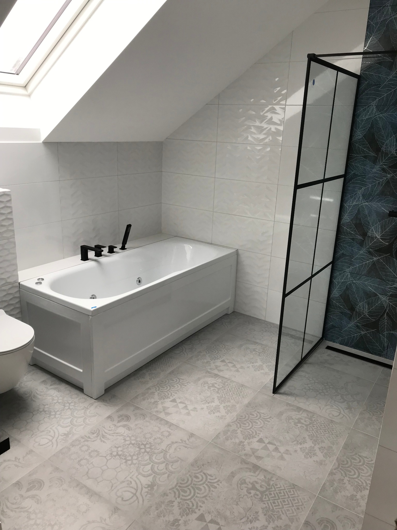 Nowoczesna łazienka na poddaszu z białą wanną z czarną armaturą, szklanym panelem prysznicowym z czarną ramą i wzorzystymi szarymi płytkami podłogowymi oraz białą ceramiką sanitarną.